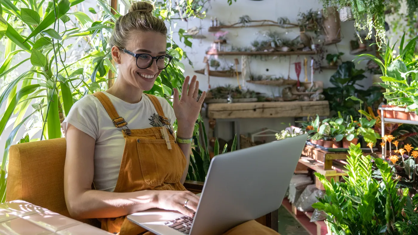 A woman wearing glasses and dungarees smiles and waves during a video call on a laptop, surrounded by plants.