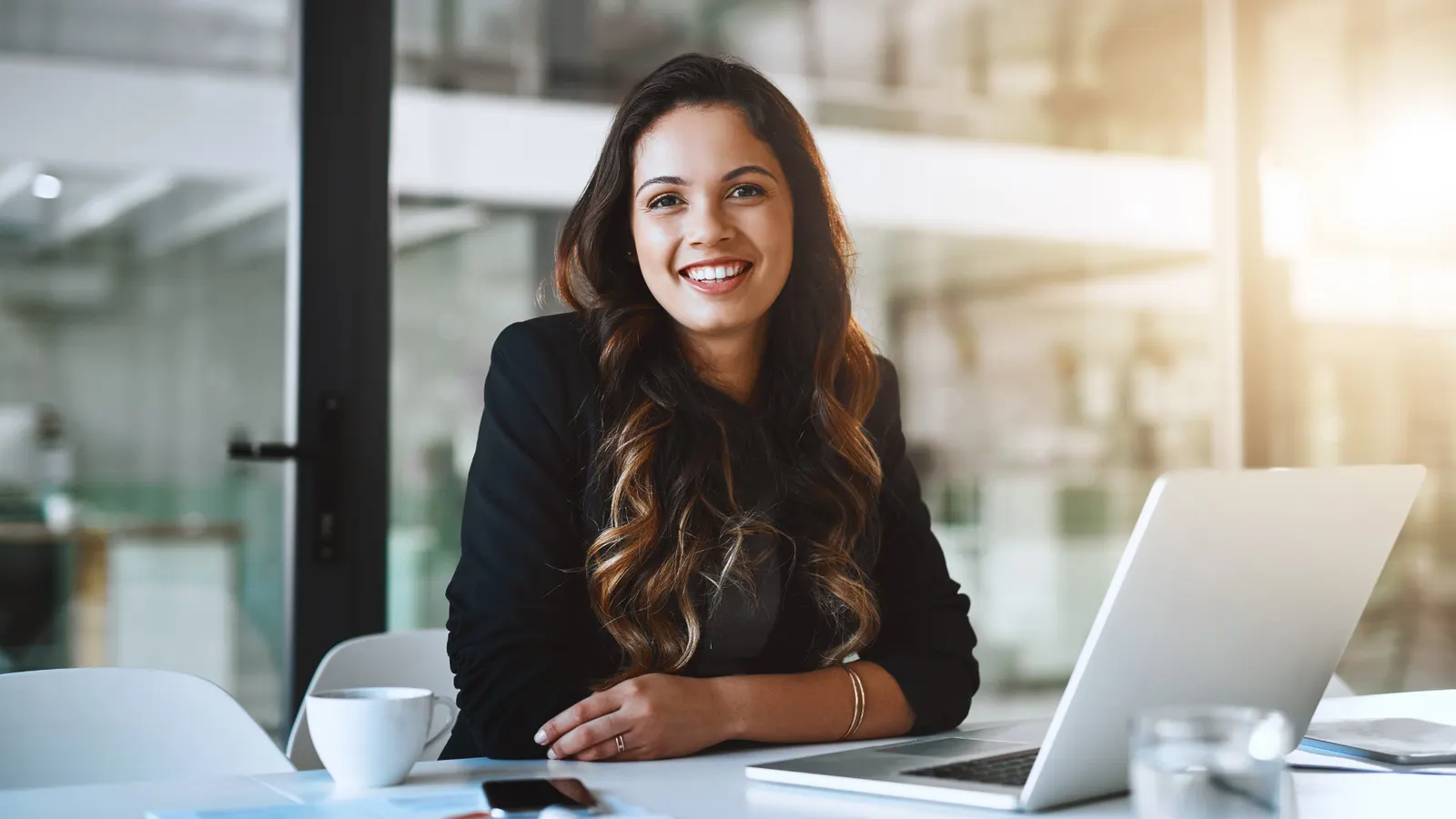 A smiling professional at a desk with a laptop in a bright office.