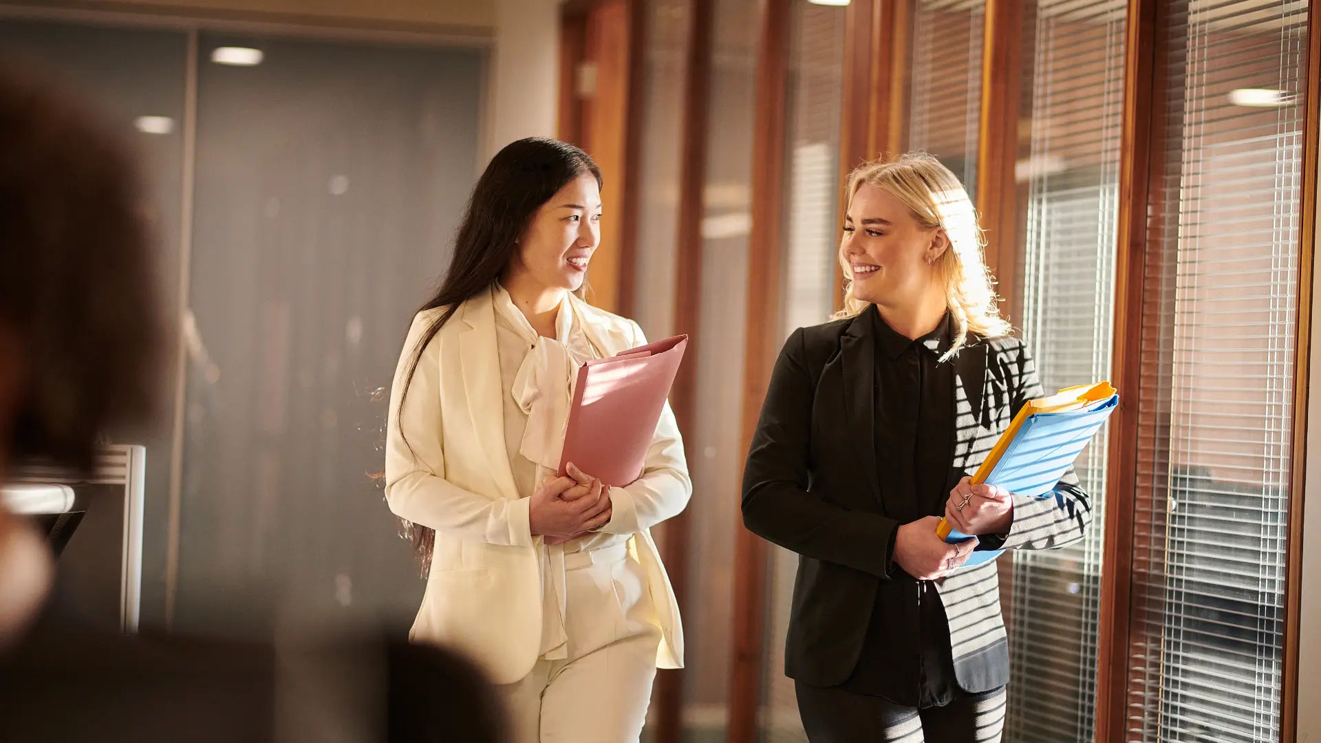 Two colleagues talking and smiling in a bright office corridor.