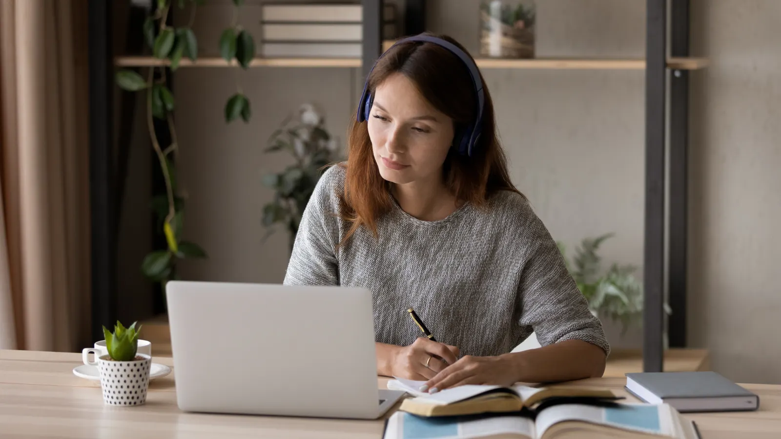 A learner wearing headphones taking notes at a laptop in a home study setting.