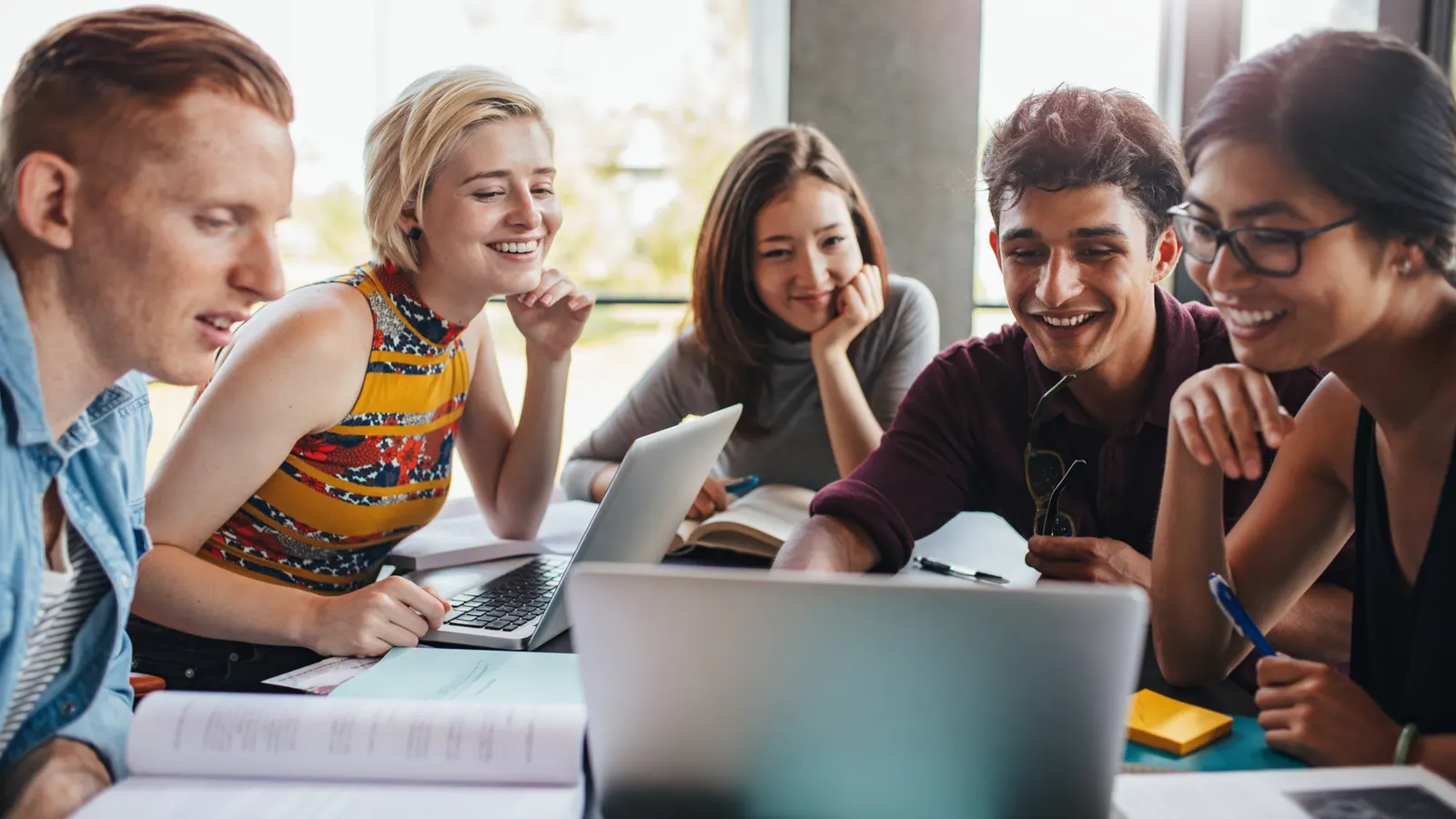 A group of five colleagues gathered around laptops, smiling and discussing together.