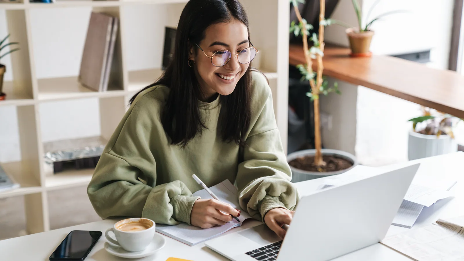 A smiling woman with glasses writing in a notebook while studying online on a laptop in a bright home workspace.