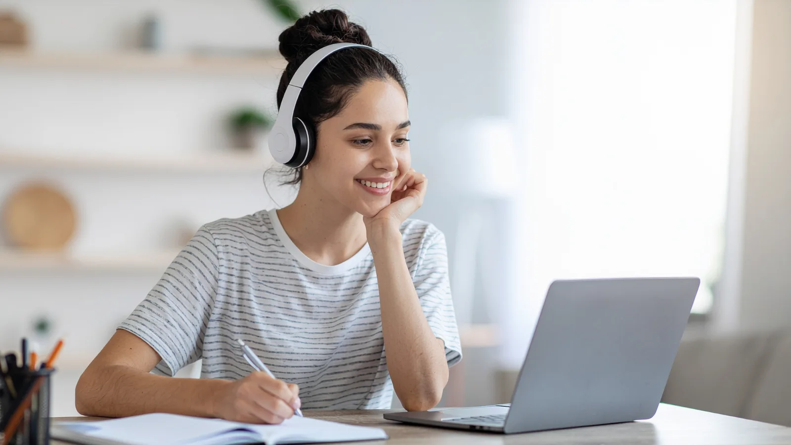 A learner wearing headphones, smiling and taking notes while studying online on a laptop.