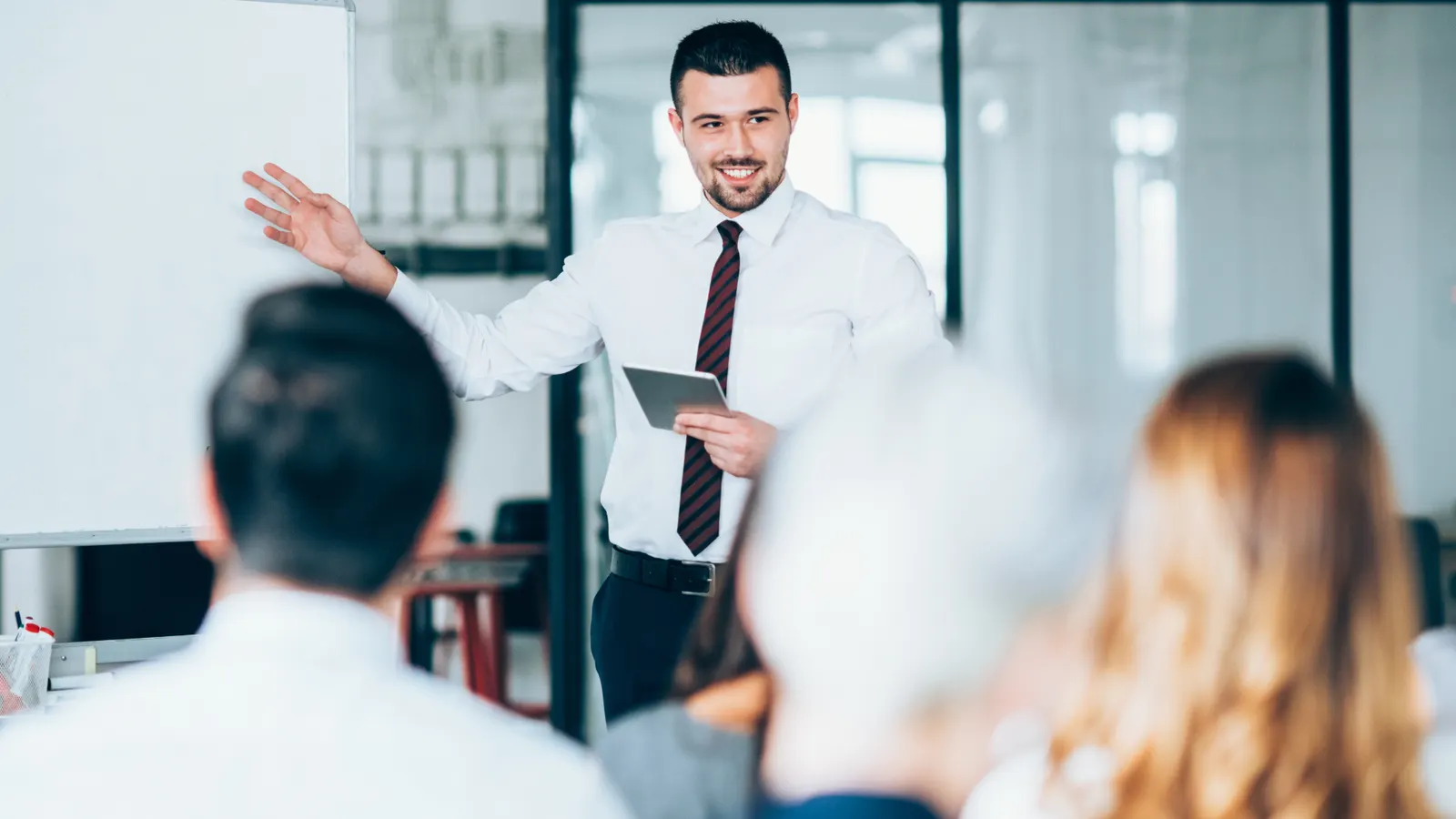 A tutor presenting to a group, gesturing with a raised hand towards a whiteboard.
